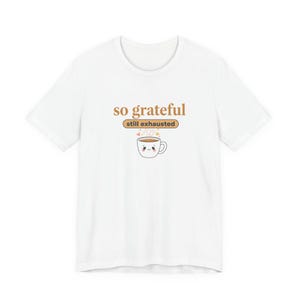 Pode incluir: Camiseta branca com a frase "so grateful" e "still exhausted". Um desenho de uma xícara de café sorrindo com vapor saindo. O design é em marrom e laranja.