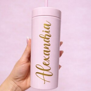 May include: A light pink tumbler with a matching lid and straw. The name "Alexandria" is written in gold cursive script vertically down the side of the tumbler. The tumbler is held in a hand against a soft pink background.