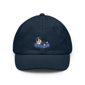May include: Navy blue baseball cap featuring an embroidered design of a black and white puppy resting on a blue pillow and holding a coffee cup. The cap has a curved brim and an adjustable strap.
