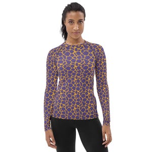 May include: A long-sleeve athletic shirt featuring a repeating giraffe-inspired pattern in shades of deep purple and golden yellow. The garment has a crew neckline and raglan sleeves, providing a fitted silhouette with a consistent print throughout.