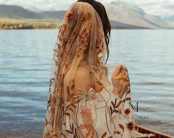 May include: A sheer, beige wedding veil with embroidered floral patterns in shades of orange, brown, and white. The veil is draped over a person standing near a lake, with mountains in the background. The text "LOVE MAI DESIGNS" is visible.