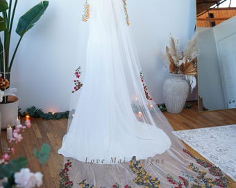 May include: A white bridal veil with a floral embroidered design in shades of red, orange, yellow, green, and blue. The veil is shown on a mannequin and has a long train.