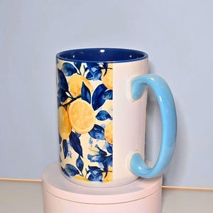 K&ouml;nnte beinhalten: Eine wei&szlig;e Keramik-Tasse mit hellblauem Griff und Innenraum. Die Tasse hat ein Zitronen- und blaues Blattdesign. Die Tasse steht auf einer rosa Oberfl&auml;che.