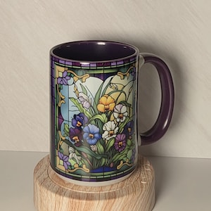 May include: A ceramic mug with a dark purple interior and handle, featuring a colorful stained glass window design on the exterior. The graphic depicts a vibrant bouquet of yellow, white, blue, and purple pansies framed by decorative gold borders.