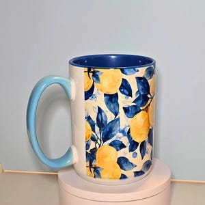 K&ouml;nnte beinhalten: Wei&szlig;er Keramikbecher mit blauem Innenraum und hellblauem Griff. Der Becher ist mit einem Muster aus gelben Zitronen und blauen Bl&auml;ttern auf cremefarbenem Hintergrund verziert. Eine stilvolle Erg&auml;nzung f&uuml;r jede K&uuml;che.