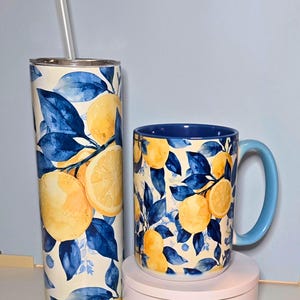 K&ouml;nnte beinhalten: Ein Becher mit Strohhalm und eine Tasse, beide mit einem Zitronen- und blaues Blattmuster. Die Tasse hat einen blauen Griff und Innenraum. Der Hintergrund ist hellblau. Die Objekte stehen auf einer wei&szlig;en Oberfl&auml;che.