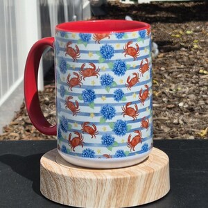 May include: A ceramic mug with a red interior and handle, featuring a repeating pattern of orange crabs, blue hydrangea flowers, and small yellow stars over a background of thin horizontal blue and white stripes on the exterior surface.
