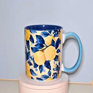 K&ouml;nnte beinhalten: Eine Keramiktasse mit einem Zitronen- und Bl&auml;ttermuster. Die Tasse hat einen hellblauen Griff und Innenraum mit einer cremefarbenen Au&szlig;enseite. Die Au&szlig;enseite ist mit gelben Zitronen und blauen Bl&auml;ttern verziert. Die Tasse steht auf einer rosa Oberfl&auml;che.