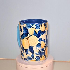 K&ouml;nnte beinhalten: Eine Keramik-Tasse mit blauem Griff und Innenraum. Die Au&szlig;enseite zeigt ein Muster aus Zitronen, Bl&auml;ttern und Zweigen in Gelb- und Blaut&ouml;nen auf cremefarbenem Hintergrund. Die Tasse ist ein K&uuml;chen- und Esszimmerartikel.