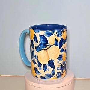 K&ouml;nnte beinhalten: Eine Keramik-Tasse mit hellblauem Henkel und Innenraum. Die Au&szlig;enseite zeigt ein Muster aus gelben Zitronen und blauen Bl&auml;ttern auf cremefarbenem Hintergrund. Die Tasse ist zylindrisch und steht auf einer rosa Oberfl&auml;che.