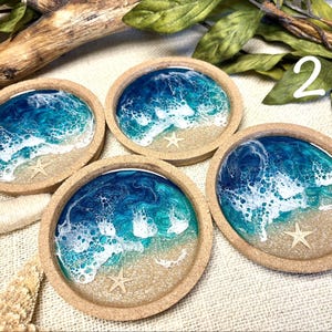 May include: Four round coasters with a cork rim and a resin inlay depicting a beach scene. Each coaster features a sandy base, blue and turquoise ocean water with white foam waves, and a small, light-colored starfish embedded in the resin.