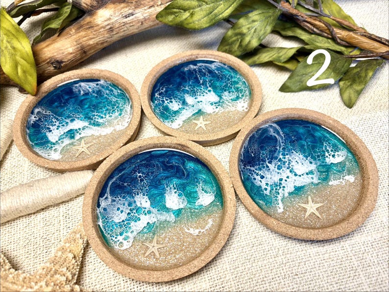 May include: Four round coasters with a cork rim and a resin inlay depicting a beach scene. Each coaster features a sandy base, blue and turquoise ocean water with white foam waves, and a small, light-colored starfish embedded in the resin.
