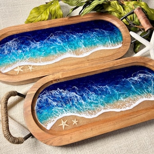 May include: Two oval wooden serving trays with a resin ocean scene inside. The resin is blue, green, and white to resemble waves crashing on a sandy beach. Each tray has small, gold, starfish accents.