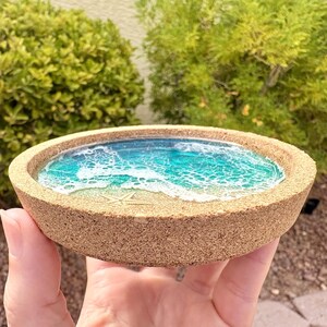 May include: A round cork coaster featuring a resin inlay that mimics a beach scene with turquoise and blue water, white foam waves, a sandy shore, and a small starfish embedded in the center of the sandy area within the circular cork frame.