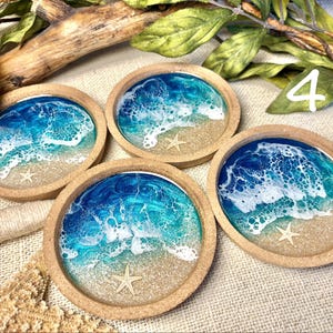 May include: Four round coasters with a raised edge, each featuring a resin art design of ocean waves crashing onto a sandy beach. The design includes shades of blue and turquoise, white sea foam, tan sand, and a small white starfish embedded in each.