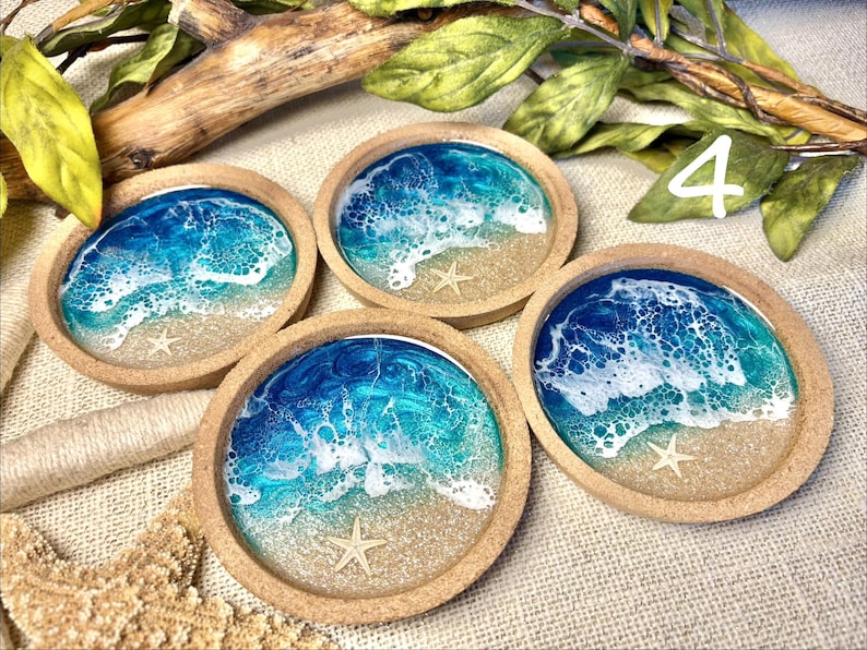 May include: Four round coasters with a raised edge, each featuring a resin art design of ocean waves crashing onto a sandy beach. The design includes shades of blue and turquoise, white sea foam, tan sand, and a small white starfish embedded in each.