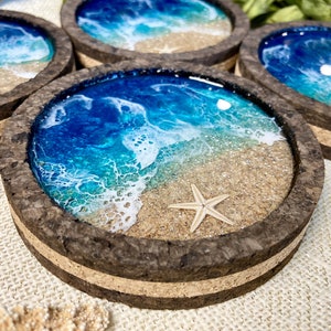 May include: A set of four circular coasters with a dark cork frame. Each coaster features a resin art design depicting a beach scene with blue ocean waves, white sea foam, tan sand, and a small embedded starfish on the sandy surface.