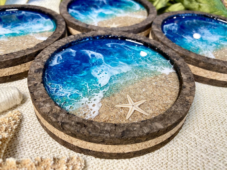 May include: A set of four circular coasters with a dark cork frame. Each coaster features a resin art design depicting a beach scene with blue ocean waves, white sea foam, tan sand, and a small embedded starfish on the sandy surface.