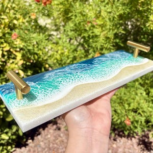 May include: A rectangular serving tray with a beach-themed design. The tray features a depiction of ocean waves in shades of blue and green, with a sandy shore. It has gold-colored handles on each end.