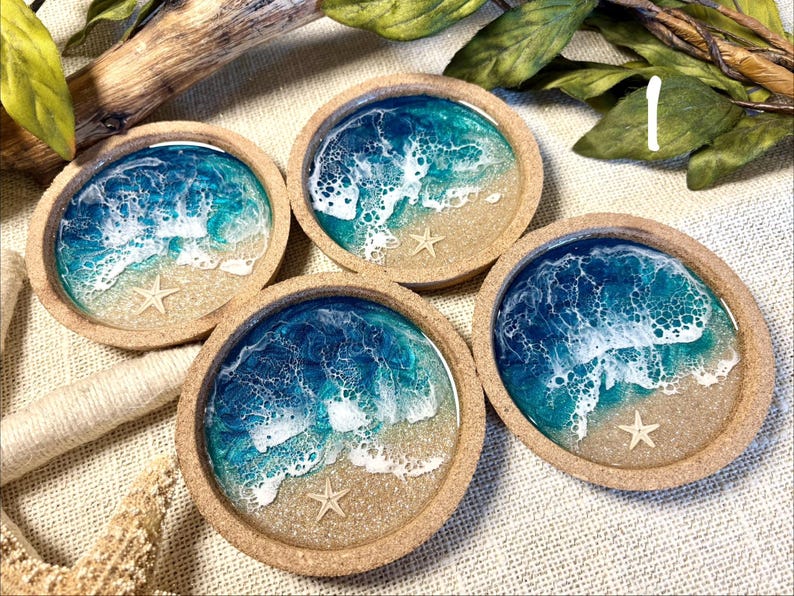 May include: Four circular coasters with a natural cork rim, featuring a resin art design that mimics a beach scene with blue and turquoise ocean waves, white sea foam, sandy beige shores, and a small embedded starfish in each piece.