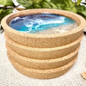 May include: A stack of four round cork coasters featuring a resin inlay that mimics a beach scene with blue and turquoise ocean waves, white sea foam, sandy beige shores, and a small white starfish embedded in the surface of the top coaster.