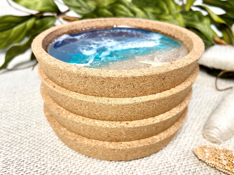 May include: A stack of four round cork coasters featuring a resin inlay that mimics a beach scene with blue and turquoise ocean waves, white sea foam, sandy beige shores, and a small white starfish embedded in the surface of the top coaster.