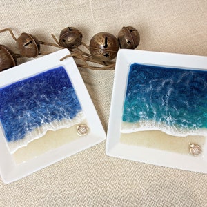 May include: Two square white ceramic dishes with a resin ocean scene inside. The dishes feature a blue and white ocean scene with a sandy beach and a small turtle figurine.