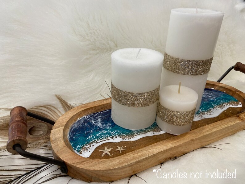 May include: Wooden tray with a resin ocean wave design and three white candles with gold glitter accents. The tray has a black metal handle.