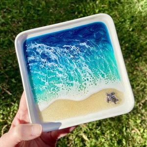 May include: A white ceramic dish with a resin ocean scene. The scene features blue and green water with white foam and a small silver turtle on the beach.