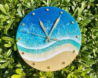 May include: Round wall clock with a beach scene design. The clock face features a blue and turquoise ocean, a sandy beach, and gold-colored hands and markers. The clock is set against a backdrop of green foliage.