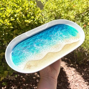 May include: An oval white tray featuring a resin art design that mimics a beach scene. The surface displays a gradient of deep blue to turquoise water with white foam waves crashing onto a textured sandy shore containing two small starfish.