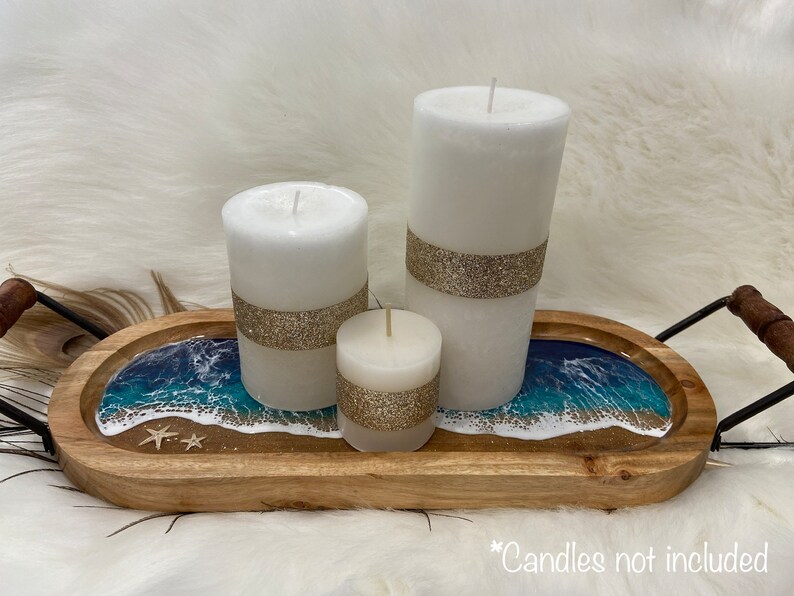 May include: A wooden tray with a resin ocean scene and three white candles with gold glitter accents. The tray has a black metal handle. *Candles not included*