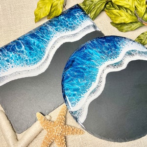 May include: Two black slate serving trays with a blue and white ocean wave design. The trays are on a beige surface with a starfish.