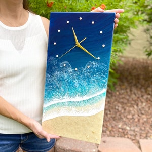 May include: A resin clock with a blue ocean wave design. The clock face is white with gold hands and white dots for the numbers. The clock is framed with a dark blue border.