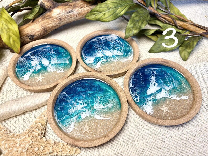 May include: Four circular coasters with a raised cork rim, each featuring a resin art design of a beach scene with blue and turquoise ocean waves, white sea foam, sandy shores, and a small embedded starfish in the center of each sandy area.