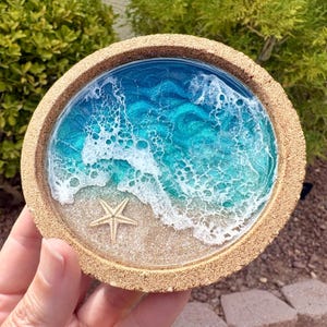 May include: A round cork coaster featuring a resin art design of a beach scene. The center displays a sandy shore with a small, light-colored starfish, transitioning into swirling shades of turquoise and deep blue ocean water with white foam.
