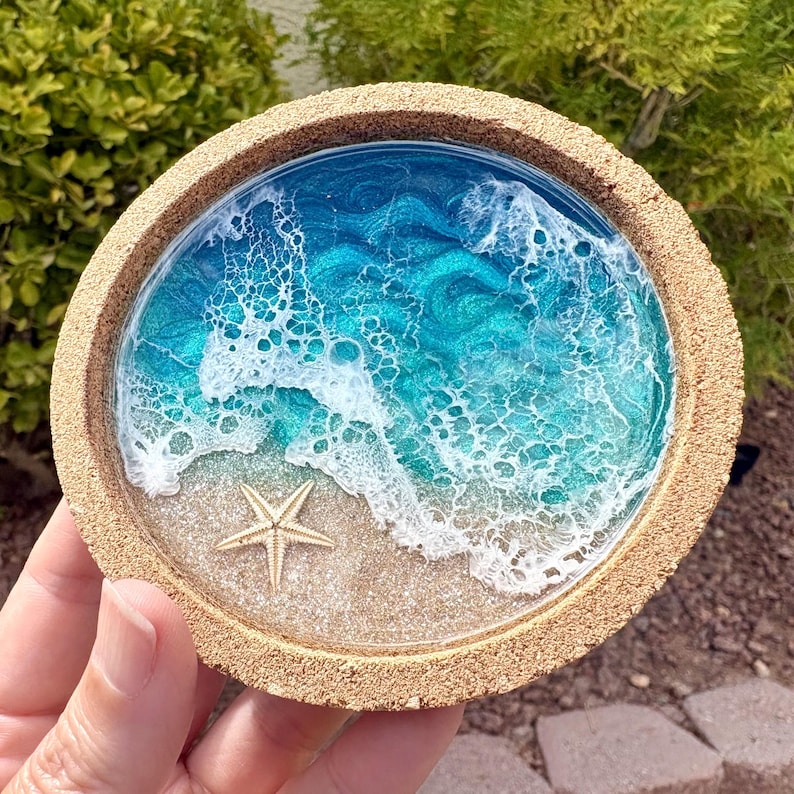 May include: A round cork coaster featuring a resin art design of a beach scene. The center displays a sandy shore with a small, light-colored starfish, transitioning into swirling shades of turquoise and deep blue ocean water with white foam.