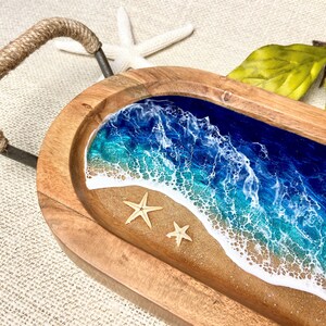 May include: A wooden serving tray with a rope handle. The tray features a resin inlay of an ocean scene with blue and green water, white foam, and a sandy beach. Two small starfish are placed on the sand.