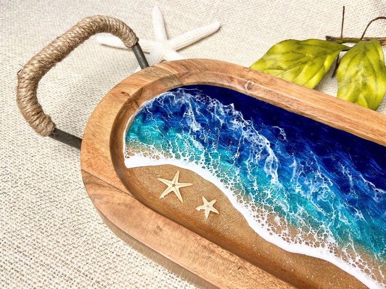 May include: A wooden serving tray with a rope handle. The tray features a resin inlay of an ocean scene with blue and green water, white foam, and a sandy beach. Two small starfish are placed on the sand.