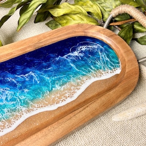 May include: A wooden serving tray with a rope handle features a blue and green ocean wave design with white foam and a sandy beach.