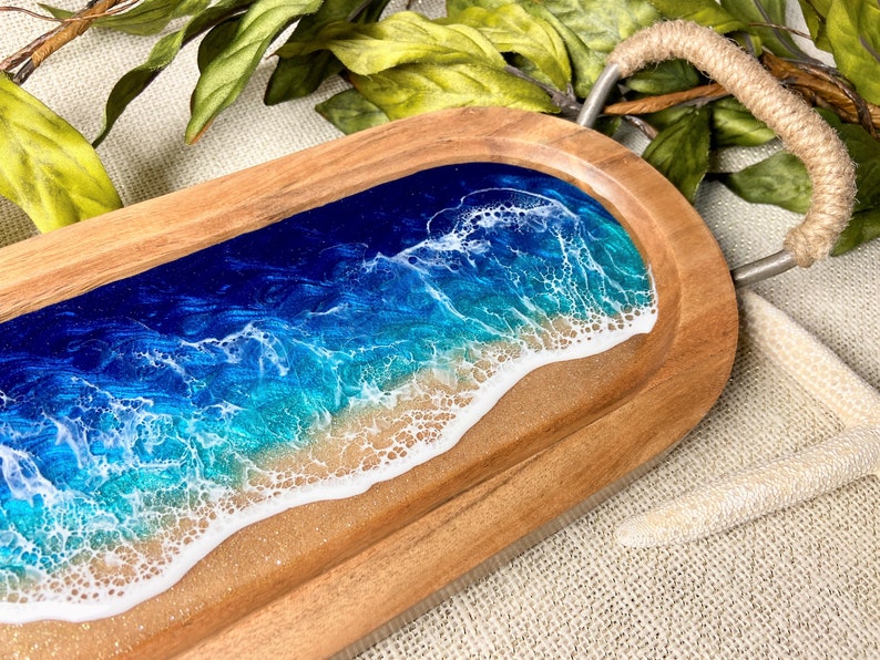 May include: A wooden serving tray with a rope handle features a blue and green ocean wave design with white foam and a sandy beach.