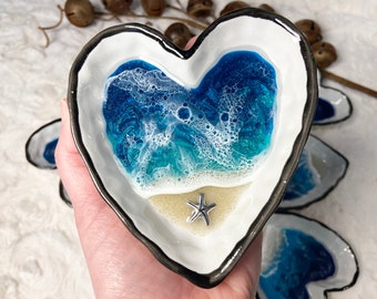 May include: A heart-shaped resin dish with a blue and white ocean wave design and a small silver starfish in the center. The dish is white with a black rim.