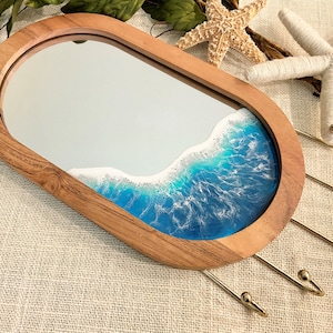 May include: An oval wooden mirror featuring a decorative resin inlay that mimics ocean waves with shades of blue and white foam. The frame is made of natural wood and includes four gold-toned metal hooks attached to the side for hanging items.