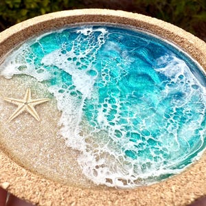 May include: A round cork coaster featuring a resin art design of a beach scene. The surface displays a sandy beige area with a five-pointed starfish, transitioning into swirling turquoise and deep blue ocean water with white foam waves.