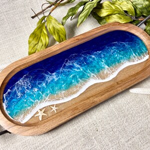 May include: A wooden serving tray with a resin inlay depicting a blue ocean wave crashing on a sandy beach. Two white starfish are embedded in the resin.