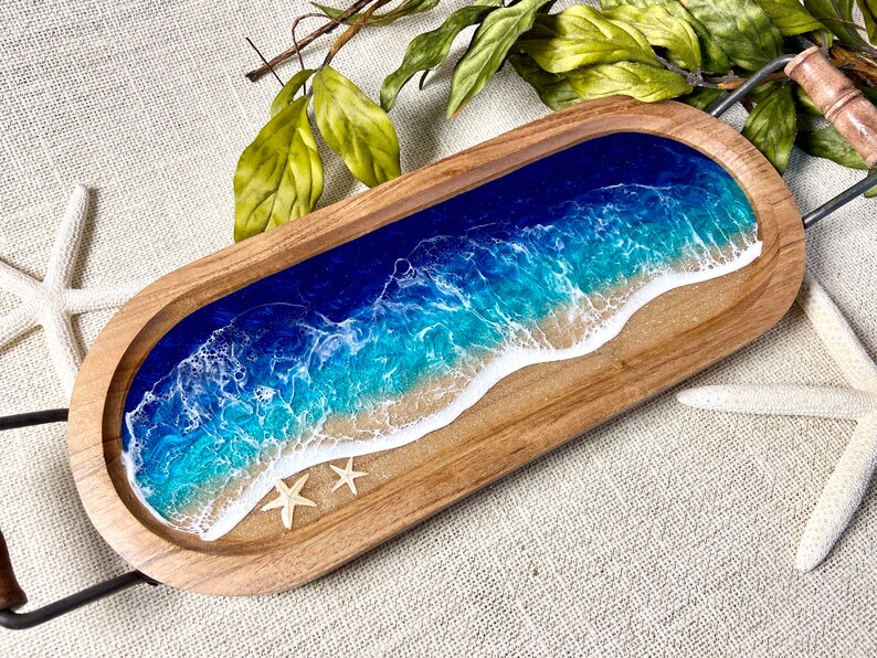 May include: A wooden serving tray with a resin inlay depicting a blue ocean wave crashing on a sandy beach. Two white starfish are embedded in the resin.
