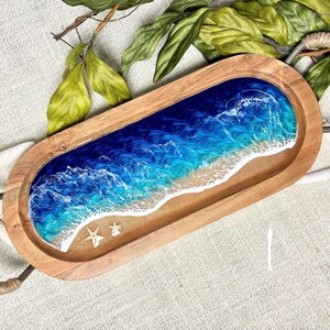 May include: A wooden serving tray with a blue and white resin ocean wave design. The tray has rope handles and is perfect for serving snacks or drinks.