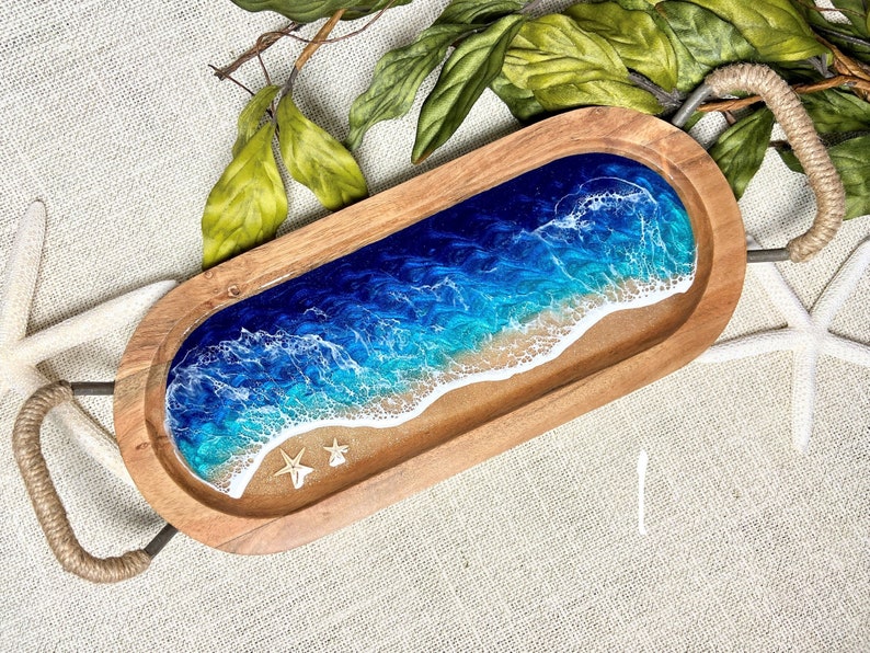 May include: A wooden serving tray with a blue and white resin ocean wave design. The tray has rope handles and is perfect for serving snacks or drinks.