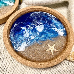 May include: A round cork coaster featuring a resin art design that mimics a beach scene. The surface displays a sandy beige base with a deep blue ocean section and white lacy foam waves. A small, light-colored starfish is embedded in the sand.