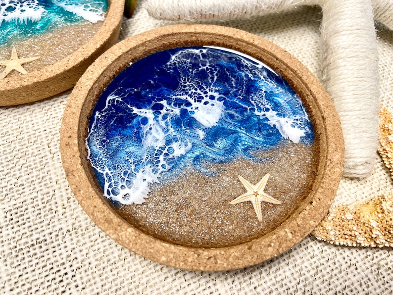 May include: A round cork coaster featuring a resin art design that mimics a beach scene. The surface displays a sandy beige base with a deep blue ocean section and white lacy foam waves. A small, light-colored starfish is embedded in the sand.